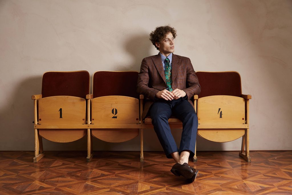 A man sitting on vintage theater seats wearing stylish formal menswear, including a patterned tie and a tailored suit, showcasing the sophistication and elegance of classic men's formal attire.