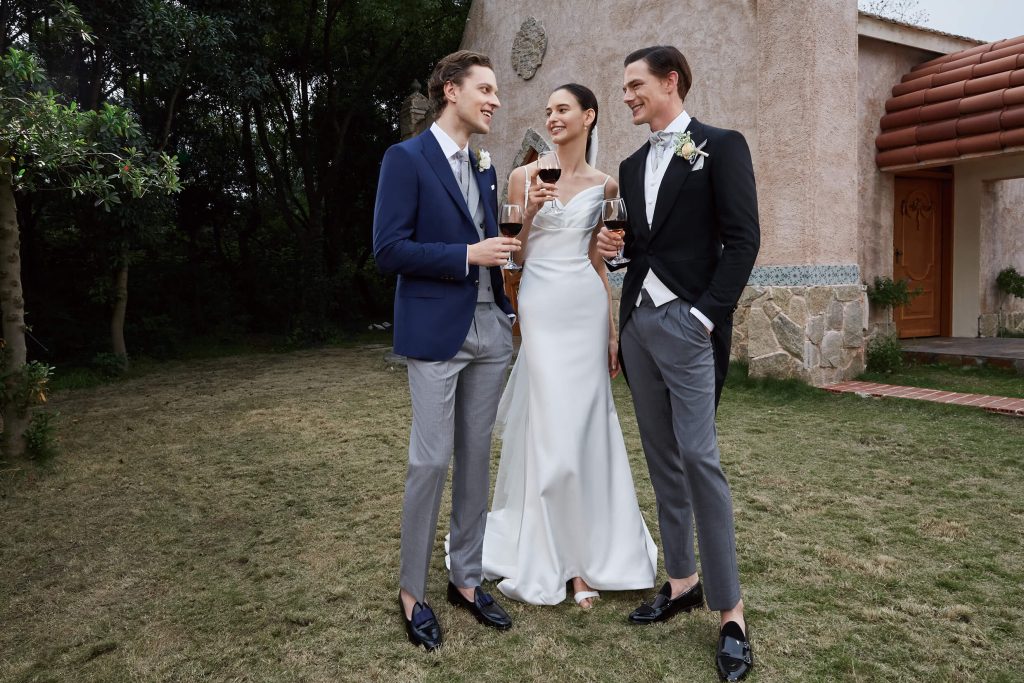 Two male London gentle men wearing a wedding dinner suit standing with a bride with a glass of wine and laughing from gisting