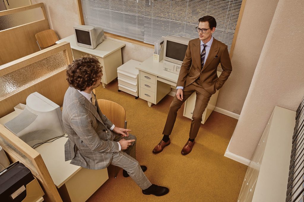 Two men in a retro office setting wearing bespoke shirts and suits, engaging in a conversation, highlighting the elegance and sophistication of custom-made fashion.