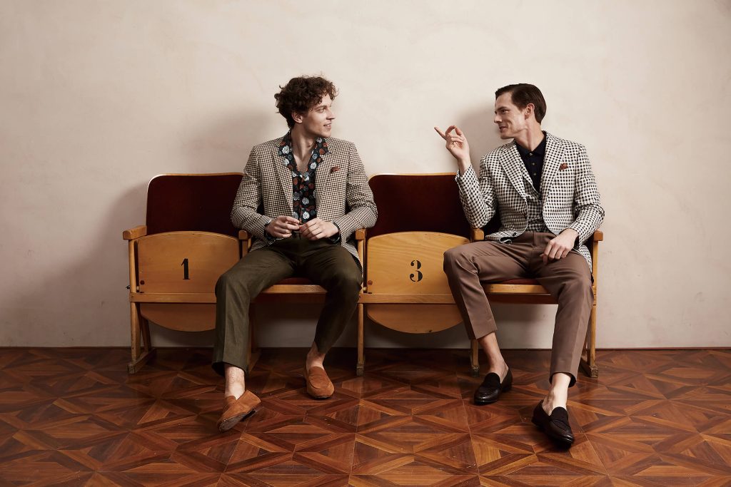 Two men sitting on vintage theater seats, wearing stylish formal menswear, including patterned blazers and tailored trousers, showcasing the elegance and versatility of classic men's formal attire.